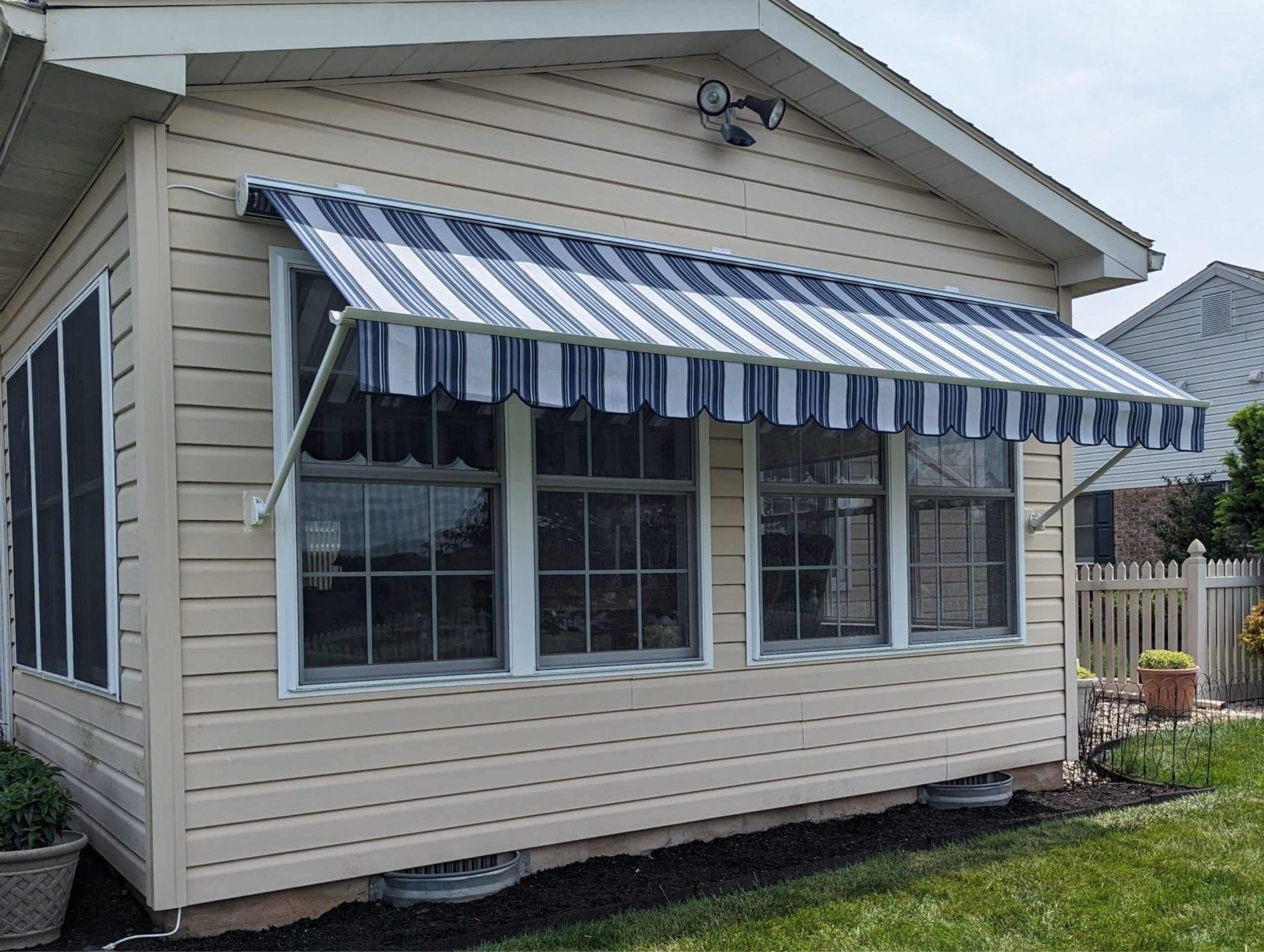 Close up of a house exterior with tan vinyl siding, white windows, and a mounted white fabric awning with dark blue and black stripes.