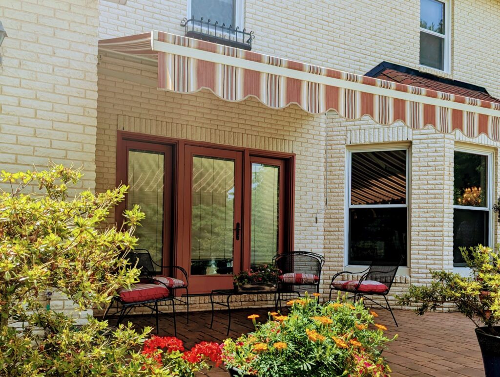 Exterior of a white brick building with a brown and glass front door, black patio set, and a red, white, gray, and black striped fabric awning.