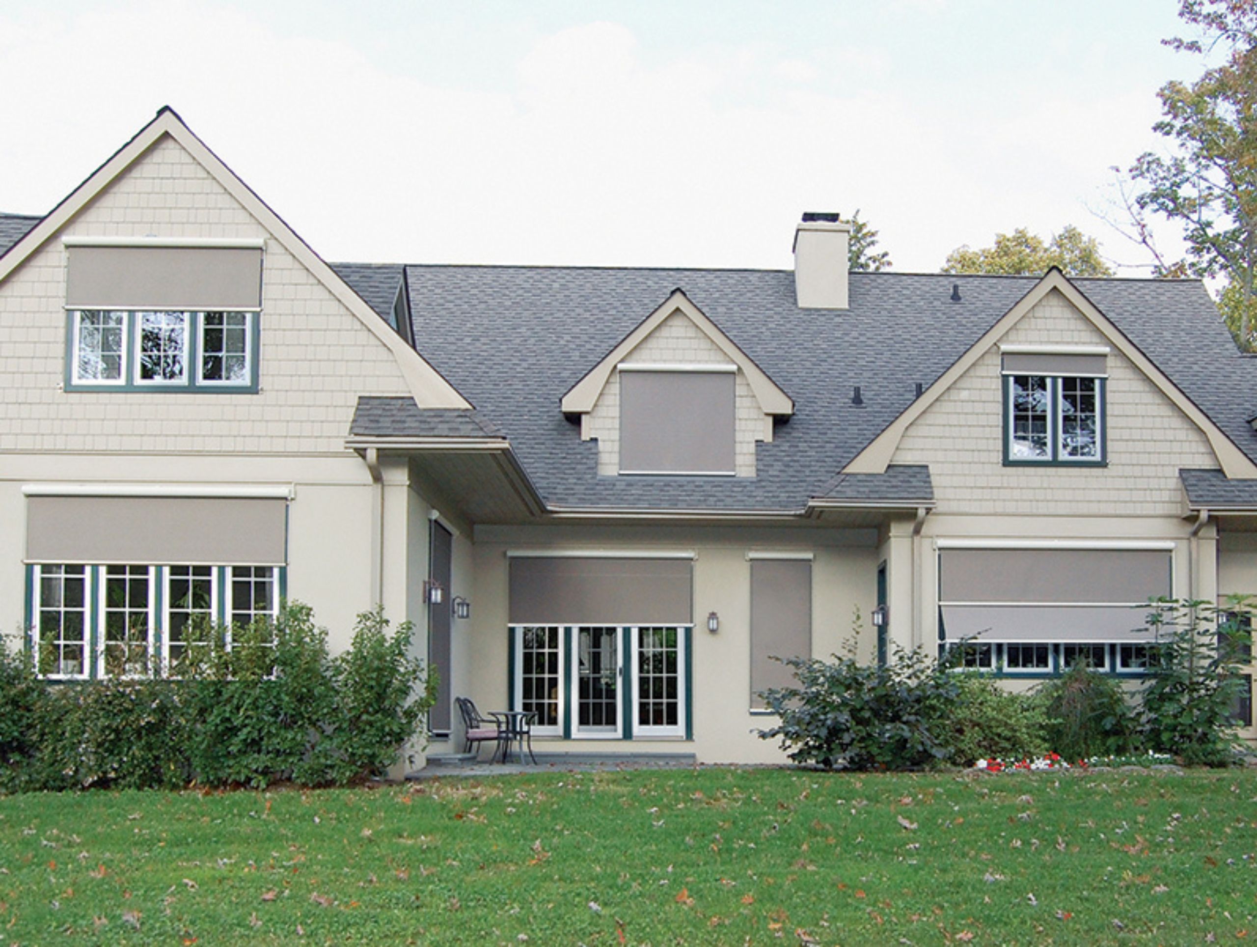 Front exterior of a tan house with a gray asphalt roof, white windows with dark green trim, and tan solar shades on windows.